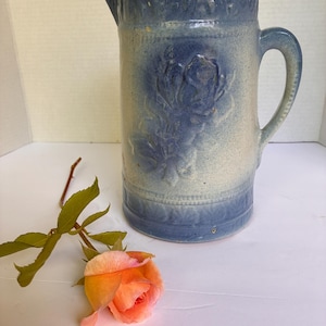 May include: A cylindrical ceramic pitcher with a blue and off-white ombre glaze, featuring a raised floral relief pattern on the front and decorative bands at the top and bottom. A single peach-colored rose with green leaves lies beside it.
