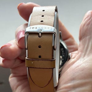 Puede incluir: Un reloj TOKYObay con una correa de cuero marr&oacute;n claro. La correa del reloj se asegura con una hebilla plateada. La esfera del reloj es visible en el lado derecho de la imagen.