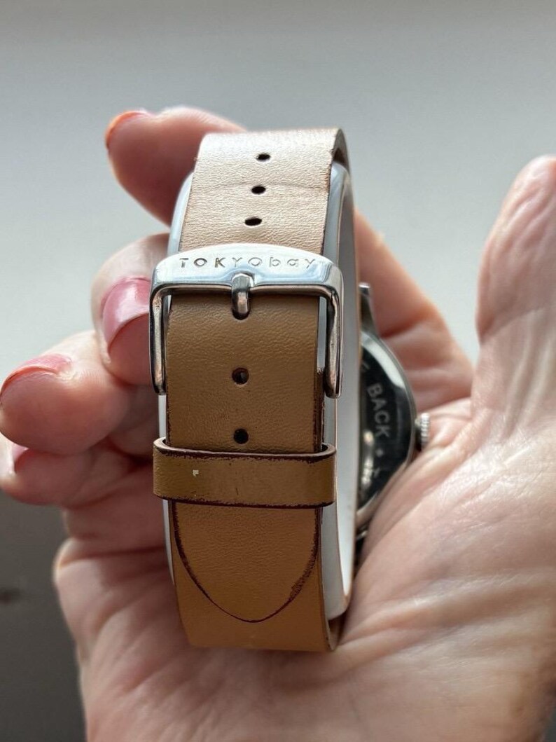 Puede incluir: Un reloj TOKYObay con una correa de cuero marr&oacute;n claro. La correa del reloj se asegura con una hebilla plateada. La esfera del reloj es visible en el lado derecho de la imagen.