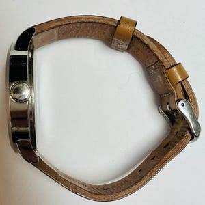 Puede incluir: Vista lateral de un reloj de pulsera con caja plateada y correa de cuero marr&oacute;n claro. La correa se sujeta con una hebilla plateada. La esfera del reloj no es visible.