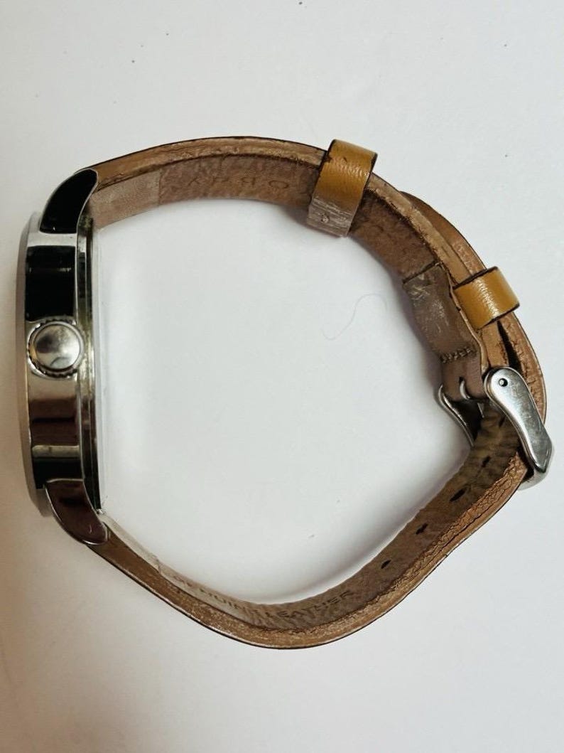 Puede incluir: Vista lateral de un reloj de pulsera con caja plateada y correa de cuero marr&oacute;n claro. La correa se sujeta con una hebilla plateada. La esfera del reloj no es visible.