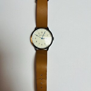 Puede incluir: Un reloj de pulsera con una correa de cuero marr&oacute;n claro y una caja plateada. La esfera del reloj es blanca con n&uacute;meros y agujas negras. El texto "TOKYO" es visible en la esfera.