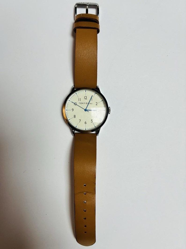 Puede incluir: Un reloj de pulsera con una correa de cuero marr&oacute;n claro y una caja plateada. La esfera del reloj es blanca con n&uacute;meros y agujas negras. El texto "TOKYO" es visible en la esfera.