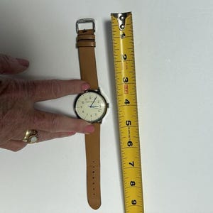 Puede incluir: Un reloj de pulsera con una correa de cuero marr&oacute;n claro y una caja de color plateado. La esfera del reloj es blanca con n&uacute;meros y manecillas negras. Una cinta m&eacute;trica amarilla est&aacute; al lado del reloj.