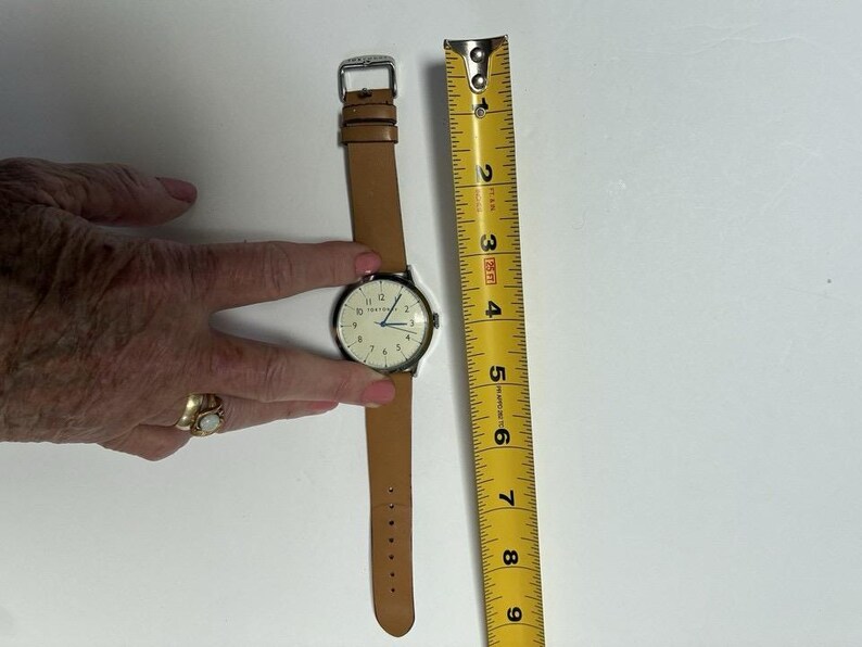 Puede incluir: Un reloj de pulsera con una correa de cuero marr&oacute;n claro y una caja de color plateado. La esfera del reloj es blanca con n&uacute;meros y manecillas negras. Una cinta m&eacute;trica amarilla est&aacute; al lado del reloj.