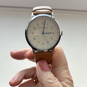 Puede incluir: Un reloj de pulsera con esfera color crema, caja plateada y correa de cuero marr&oacute;n. La esfera del reloj muestra n&uacute;meros negros y el nombre de la marca "TOKYObay". El reloj es sostenido por una persona.