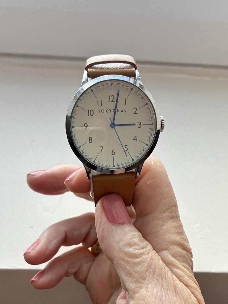 Puede incluir: Un reloj de pulsera con esfera color crema, caja plateada y correa de cuero marr&oacute;n. La esfera del reloj muestra n&uacute;meros negros y el nombre de la marca "TOKYObay". El reloj es sostenido por una persona.