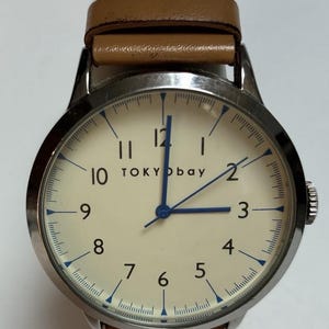 Puede incluir: Un reloj de pulsera con esfera color crema, manecillas azules y correa de cuero marr&oacute;n. La esfera del reloj muestra la marca "TOKYObay" y presenta marcadores azules. El reloj tiene una caja de metal plateado.