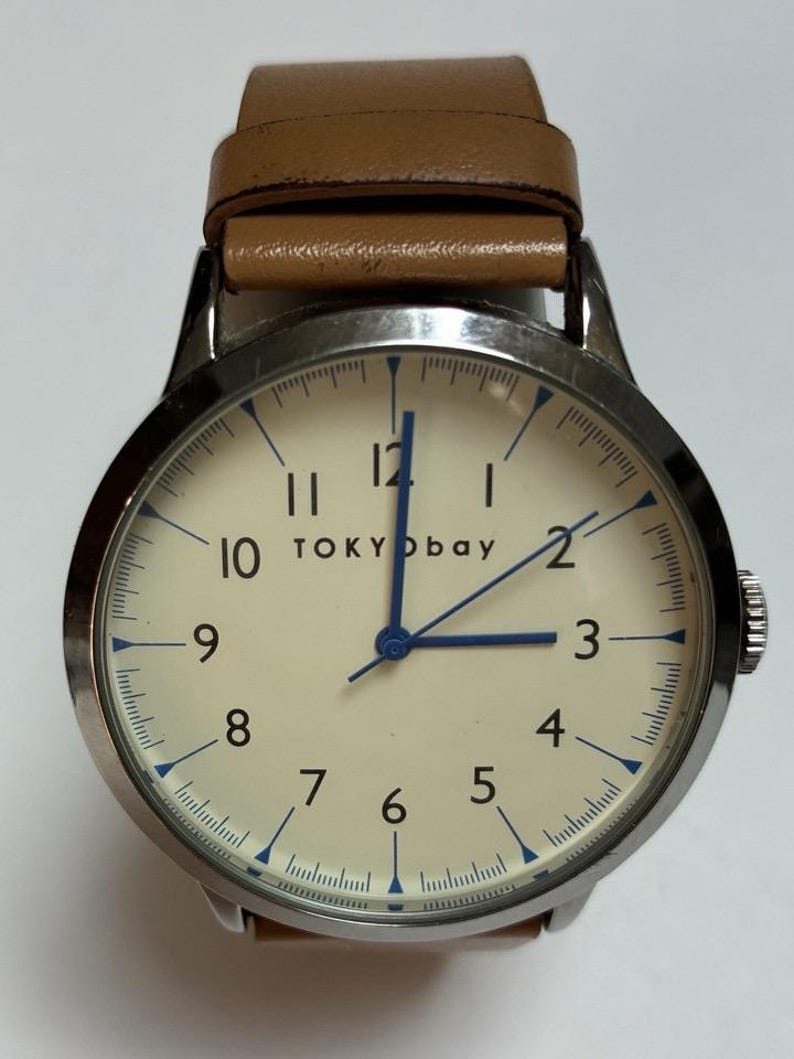 Puede incluir: Un reloj de pulsera con esfera color crema, manecillas azules y correa de cuero marr&oacute;n. La esfera del reloj muestra la marca "TOKYObay" y presenta marcadores azules. El reloj tiene una caja de metal plateado.