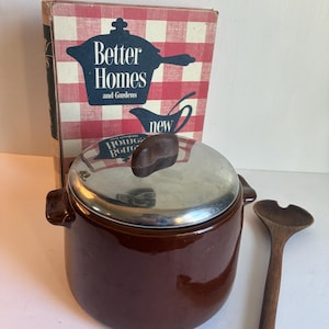 May include: A brown ceramic casserole dish with a silver lid and a dark brown handle. A wooden spoon rests beside the dish. A vintage cookbook titled "Better Homes and Gardens" is in the background.