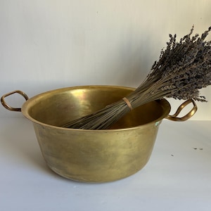 May include: A vintage brass bowl with two handles, filled with a bundle of dried lavender. The bowl has a warm, golden hue and a rounded shape. The lavender is tied with a rubber band.