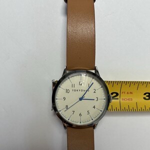 Puede incluir: Un reloj de pulsera con esfera clara y correa de cuero marr&oacute;n. La esfera del reloj tiene n&uacute;meros negros y agujas azules. El reloj tiene aproximadamente 5 cm de di&aacute;metro.