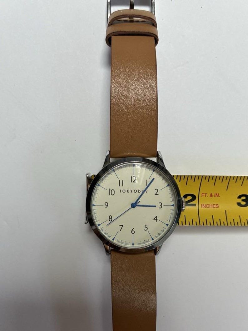 Puede incluir: Un reloj de pulsera con esfera clara y correa de cuero marr&oacute;n. La esfera del reloj tiene n&uacute;meros negros y agujas azules. El reloj tiene aproximadamente 5 cm de di&aacute;metro.