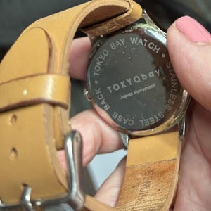 Puede incluir: Primer plano de un reloj Tokyo Bay con una correa de cuero marr&oacute;n claro. La esfera del reloj no es visible, pero la parte trasera del reloj es visible, con las palabras "TOKYO BAY WATCH" y "STAINLESS STEEL" impresas en ella.