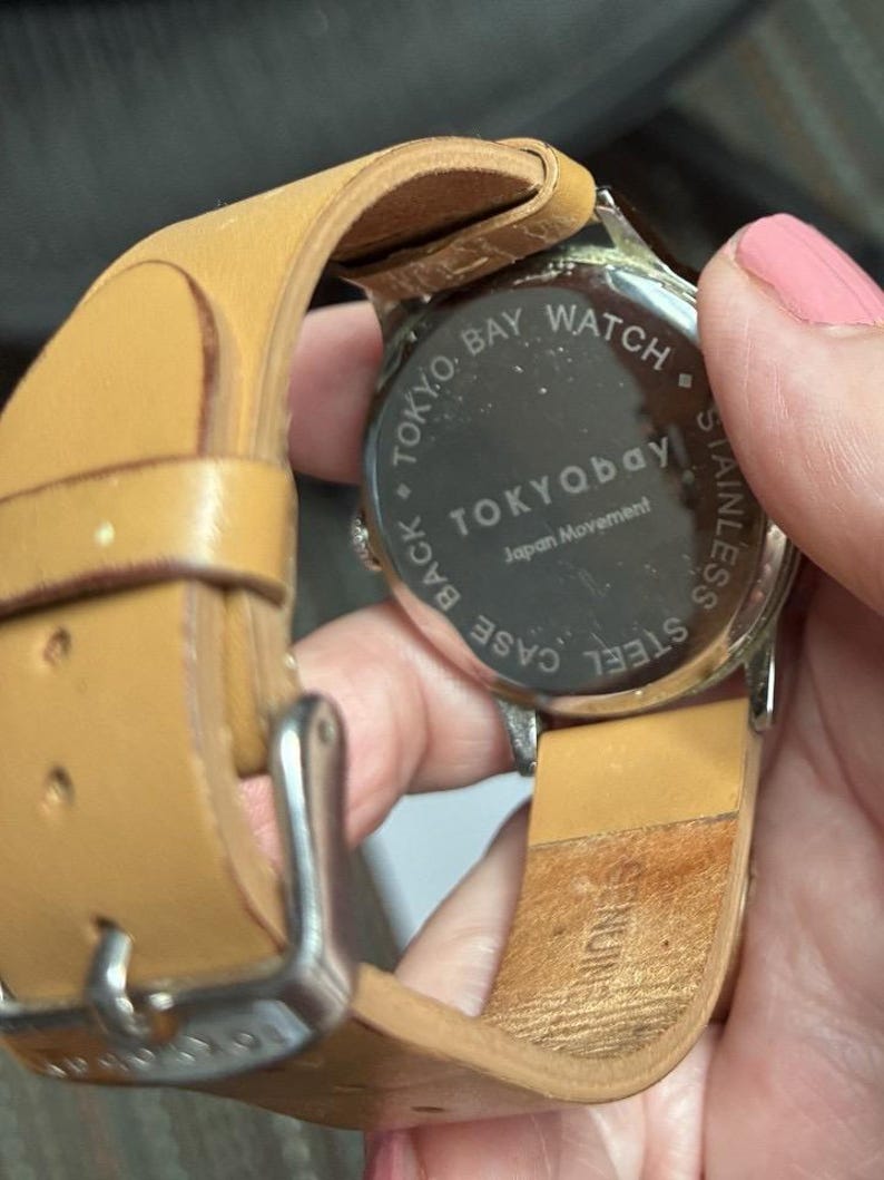 Puede incluir: Primer plano de un reloj Tokyo Bay con una correa de cuero marr&oacute;n claro. La esfera del reloj no es visible, pero la parte trasera del reloj es visible, con las palabras "TOKYO BAY WATCH" y "STAINLESS STEEL" impresas en ella.