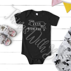 May include: Black baby onesie with the text "CUTE WITH THE 'E'" in white. The onesie is surrounded by baby shoes and a patterned blanket. The onesie is a one-piece garment with short sleeves.