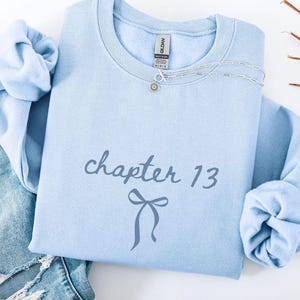 May include: A light blue sweatshirt with the text "chapter 13" and a bow graphic. A silver necklace is draped on the sweatshirt. The sweatshirt is paired with light blue jeans. The words "light blue" are at the bottom.