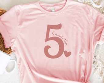 May include: A pink t-shirt with a large, rose gold number "5" and the name "Samantha" written vertically next to it. A small heart is to the right of the number.