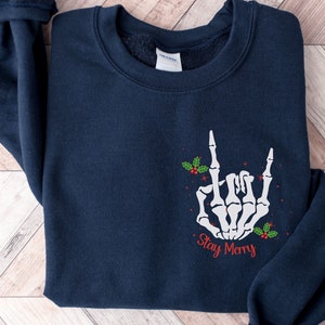 May include: A navy blue sweatshirt with a white skeleton hand making the rock and roll sign. The hand is decorated with red holly berries and green leaves. The text "Stay Merry" is printed below the hand in red.