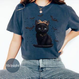 May include: A navy blue t-shirt featuring a black cat illustration with a bow and a collar, surrounded by bats and stars. The word "Midnight" is printed on a circular label on the shirt.