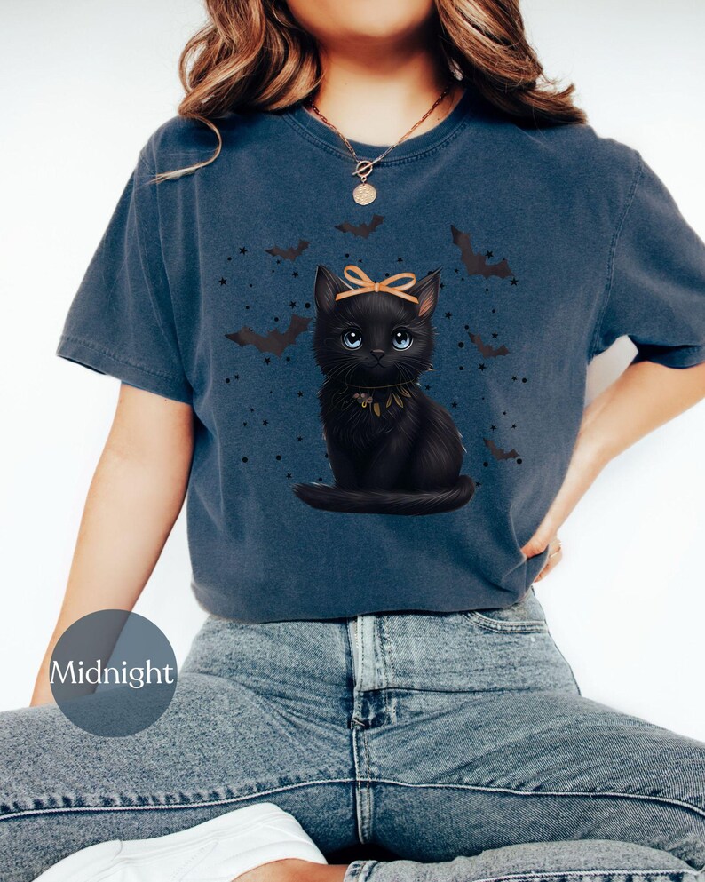 May include: A navy blue t-shirt featuring a black cat illustration with a bow and a collar, surrounded by bats and stars. The word "Midnight" is printed on a circular label on the shirt.
