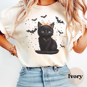 May include: Ivory-colored sweatshirt featuring a black cat illustration with a peach bow and blue eyes. The design includes bats and stars, creating a Halloween theme. The word "Ivory" is printed on the bottom right.
