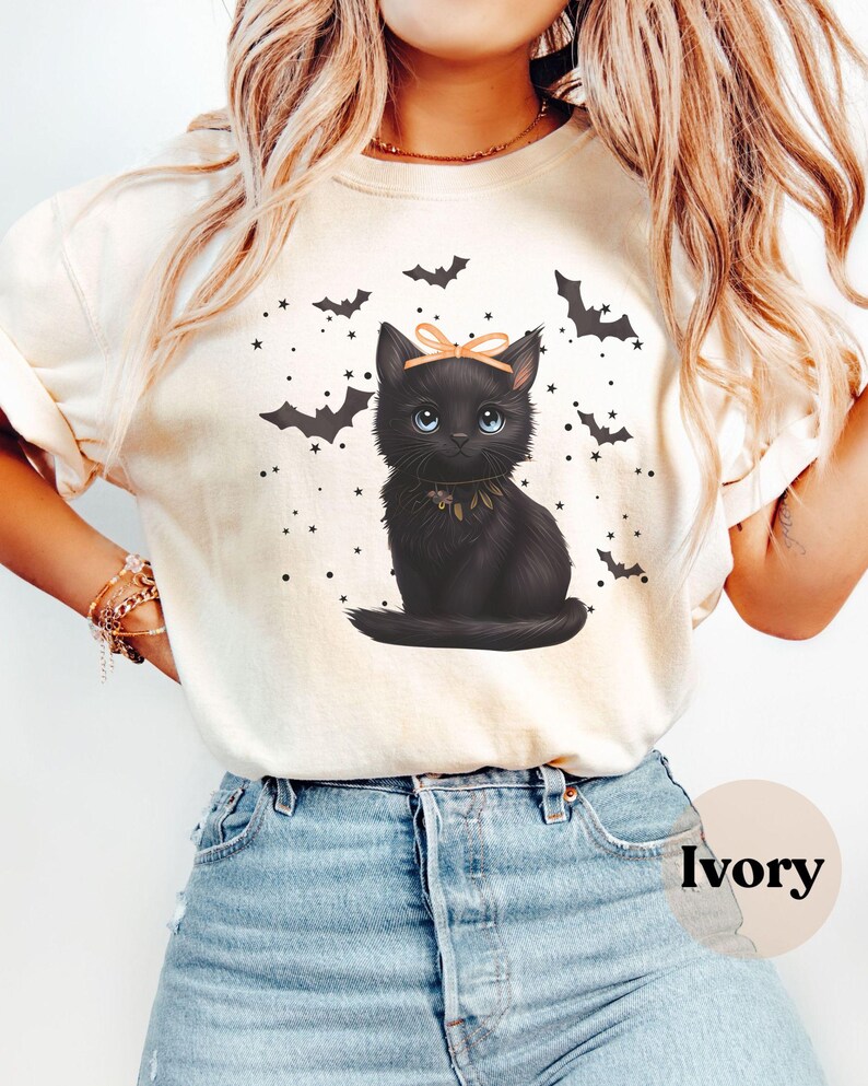 May include: Ivory-colored sweatshirt featuring a black cat illustration with a peach bow and blue eyes. The design includes bats and stars, creating a Halloween theme. The word "Ivory" is printed on the bottom right.