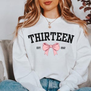 May include: White sweatshirt with black text that reads "THIRTEEN EST. 2012" and a pink bow.