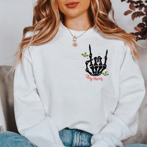 May include: White sweatshirt with a black skeleton hand making a rock and roll sign. The hand is surrounded by red berries and green holly leaves. The text "Stay Merry" is printed below the hand.