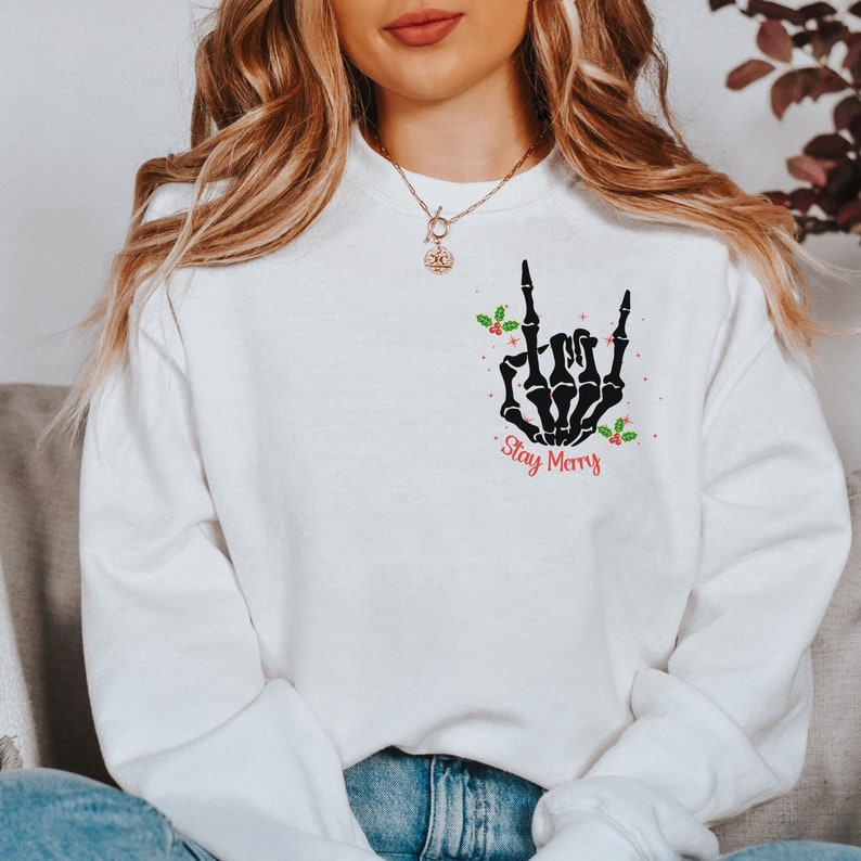 May include: White sweatshirt with a black skeleton hand making a rock and roll sign. The hand is surrounded by red berries and green holly leaves. The text "Stay Merry" is printed below the hand.