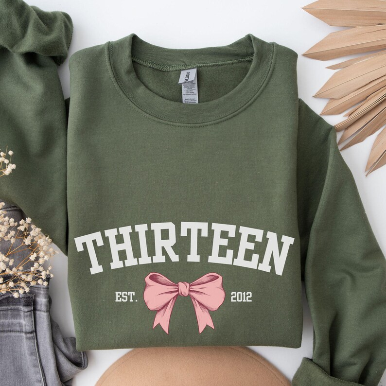 May include: Olive green sweatshirt with white text that reads "THIRTEEN EST. 2012" and a pink bow.