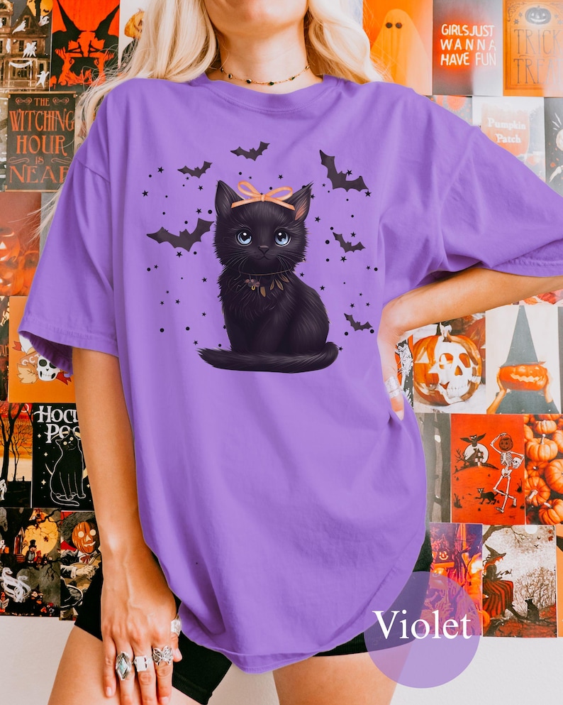 May include: A violet-colored t-shirt featuring a black cat illustration with a pink bow, surrounded by bats and stars. The text "Violet" is at the bottom. Halloween-themed apparel.