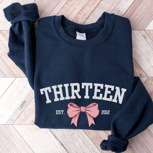 May include: Navy blue sweatshirt with white text that reads "THIRTEEN EST. 2012" and a pink bow.