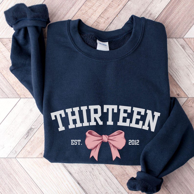 May include: Navy blue sweatshirt with white text that reads "THIRTEEN EST. 2012" and a pink bow.
