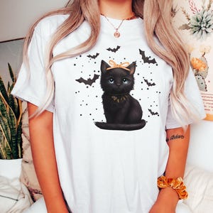 May include: White t-shirt featuring a black cat illustration with a bow, surrounded by bats and stars. The cat has blue eyes and a decorative collar. A Halloween-themed graphic tee.