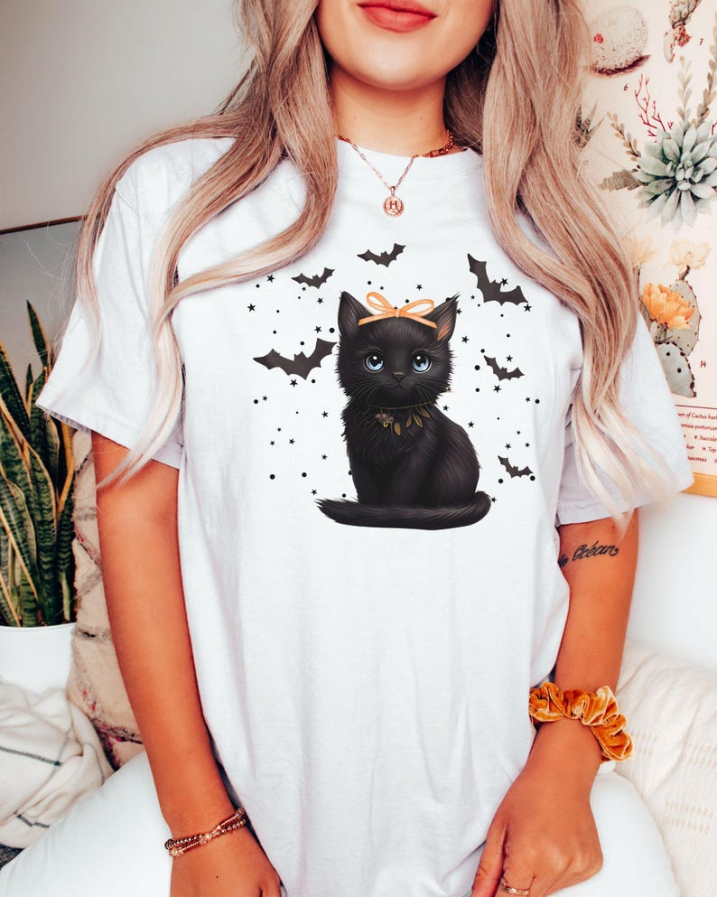 May include: White t-shirt featuring a black cat illustration with a bow, surrounded by bats and stars. The cat has blue eyes and a decorative collar. A Halloween-themed graphic tee.