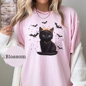 May include: Pink t-shirt featuring a black cat illustration with a bow and bat graphics. The cat has blue eyes and a decorative collar. The word "Blossom" is printed on a pink circle.