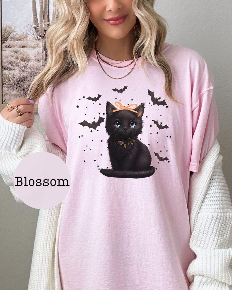 May include: Pink t-shirt featuring a black cat illustration with a bow and bat graphics. The cat has blue eyes and a decorative collar. The word "Blossom" is printed on a pink circle.