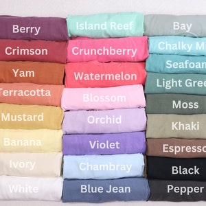 May include: A collection of folded t-shirts in various colors, including Berry, Crimson, Yam, Terracotta, Mustard, Banana, Ivory, White, Island Reef, Crunchberry, Watermelon, Blossom, Orchid, Violet, Chambray, and Blue Jean. Each shirt has a color name printed on it.