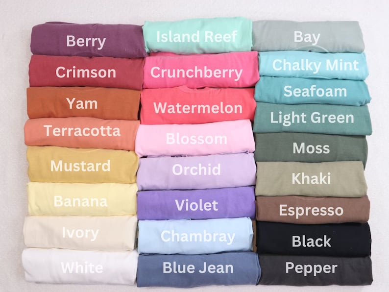May include: A collection of folded t-shirts in various colors, including Berry, Crimson, Yam, Terracotta, Mustard, Banana, Ivory, White, Island Reef, Crunchberry, Watermelon, Blossom, Orchid, Violet, Chambray, and Blue Jean. Each shirt has a color name printed on it.
