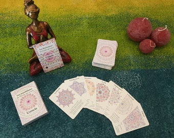 May include: A deck of oracle cards with colorful mandala designs. The cards are fanned out on a green and blue rug. The box for the cards is white with a pink mandala design and the text "Les Mandalas de l'Étoile qui Sommet L'Oracle".