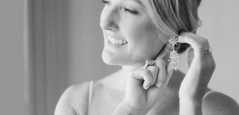 May include: A woman is putting on a pair of large, sparkly earrings. The earrings are silver and have a floral design. She is wearing a white dress and has her hair pulled back.
