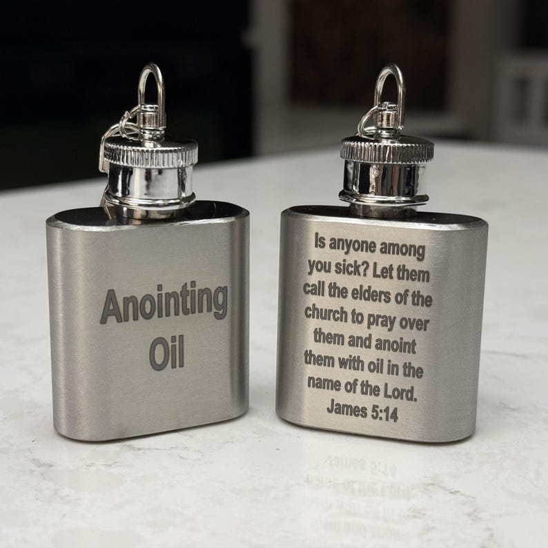 May include: Two silver metal flasks with a hinged lid and a loop for hanging. One flask is engraved with the words "Anointing Oil". The other flask is engraved with a Bible verse from James 5:14, "Is anyone among you sick? Let them call the elders of the church to pray over them and anoint them with oil in the name of the Lord. James 5:14".