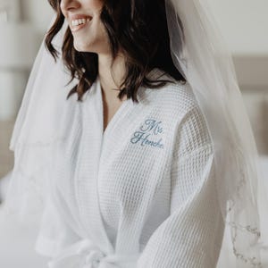 Könnte beinhalten: Weißer Waffel-Bademantel mit blau gesticktem Schriftzug "Mrs. Henche" auf der Brust. Auf dem Ärmel steht das Datum "08.08.25". Ein durchsichtiger Schleier ist über die Schultern drapiert. Der Bademantel ist an der Taille gebunden.