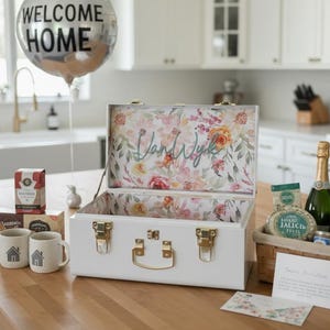May include: A white suitcase with gold hardware and a floral interior sits open on a wooden countertop. A silver balloon with "WELCOME HOME" text floats above. The scene includes mugs, tea, and a basket with food items.
