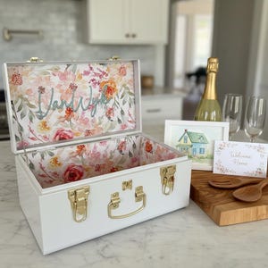 May include: White decorative box with gold hardware and a floral interior. The inside lid features the name "Vand Wyk" in cursive. A watercolor painting of a house and a "Welcome Home" card are on a wooden cutting board.