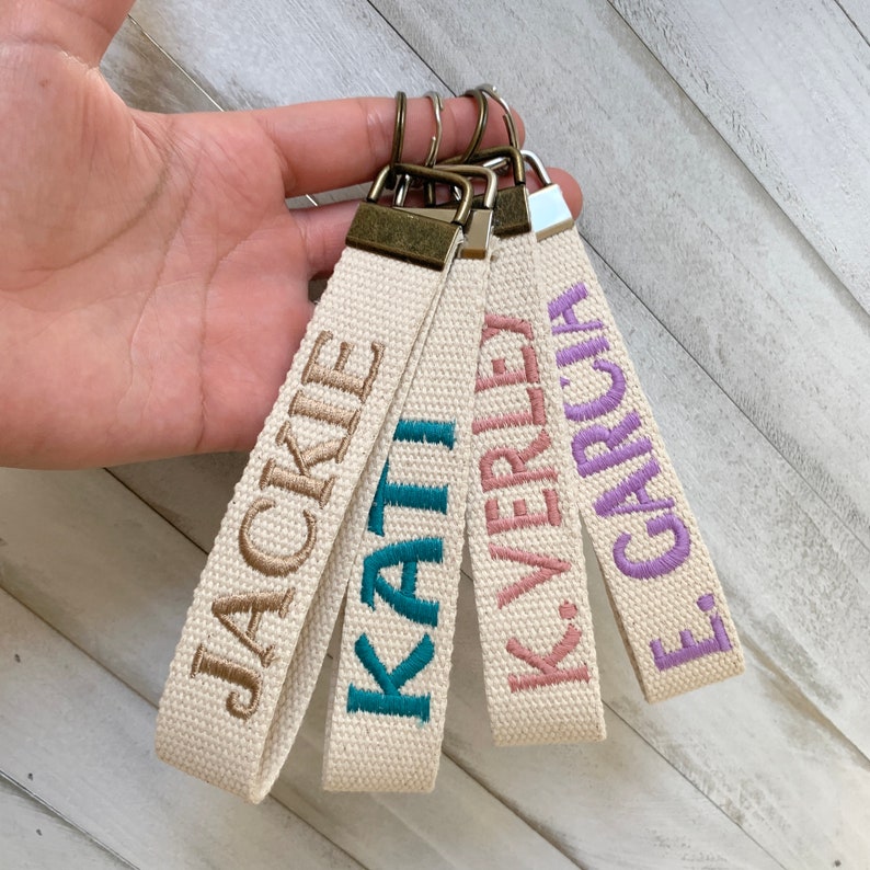 May include: Four beige fabric keychains with embroidered names in various colors: Jackie, Kati, K. Verley, and E. Garcia. Each keychain has a metal clasp and a loop for easy attachment to keys or bags. The names are in brown, teal, pink, and purple.