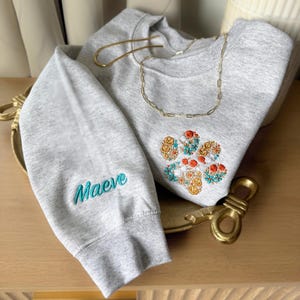 May include: A light gray crewneck sweatshirt featuring an embroidered floral paw print design on the chest and the name Maeve embroidered in teal on the sleeve. A gold-toned chain necklace rests on the neckline of the garment.