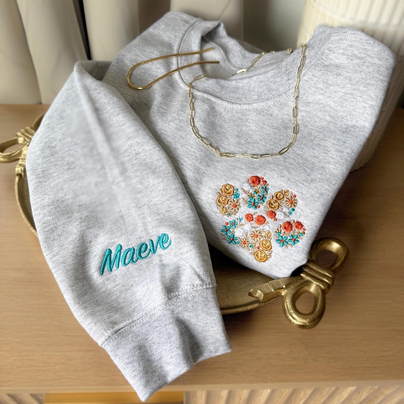 May include: A light gray crewneck sweatshirt featuring an embroidered floral paw print design on the chest and the name Maeve embroidered in teal on the sleeve. A gold-toned chain necklace rests on the neckline of the garment.