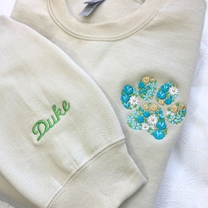 May include: A light beige sweatshirt with a green embroidered name "Duke" on the left sleeve and a blue and white floral paw print on the right side.